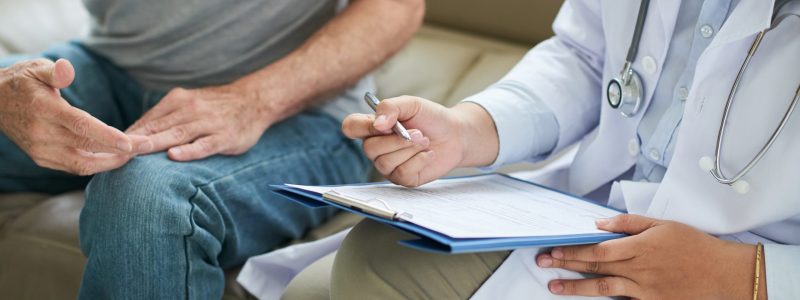 Crop shot of senior man sitting on sofa with doctor during home visit taking notes on clipboard